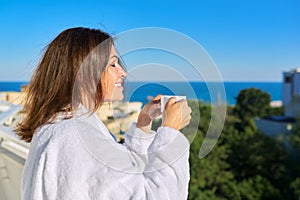 Happy mature female in white bathrobe on balcony of hotel in seaside resort town