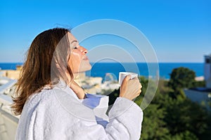 Happy mature female in white bathrobe on balcony of hotel in seaside resort town