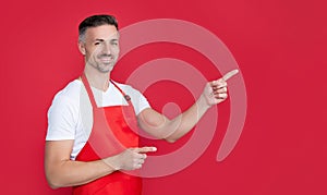happy mature householding man in apron on red background. pointing finger