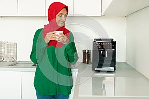 Happy mature islamic woman enjoying fresh aromatic coffee near modern machine in kitchen interior