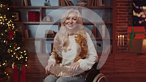Happy Mature Lady in Festive Mood Sitting in Wheelchair in Decorated House, Wearing Tinsel. Senior Smiling Woman Posing