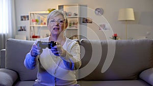 Happy mature lady playing video game on console, sitting home on sofa, leisure