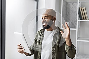 Happy mature latin man with beard in wireless earphones looking at tablet and waving hand at webcamera in home office