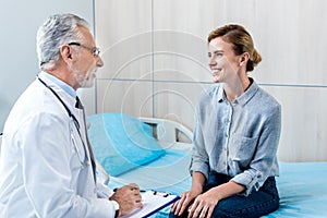 happy mature male doctor writing in clipboard near female patient