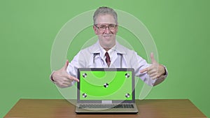 Happy mature man doctor showing laptop and giving thumbs up against wooden table