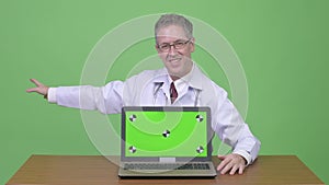 Happy mature man doctor showing laptop while presenting something against wooden table