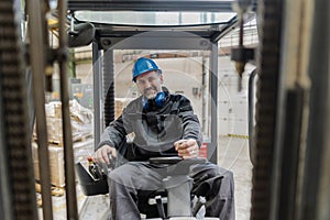 Happy mature man fork lift truck driver lifting pallet in storage warehouse and looking at camera.