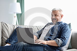 Happy mature man using laptop while resting on sofa