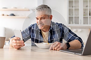 Happy mature man using smartphone while having breakfast in kitchen table at home, copy space