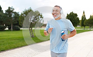 Happy mature man wearing headphones with water bottle in hand jogging outside in city park