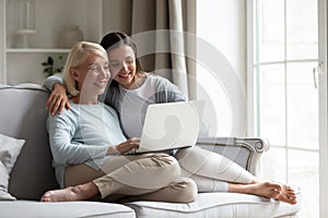 Happy mature mother and daughter sitting on couch, using laptop