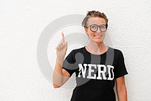 Happy mature nerd woman wearing big eyeglasses and standing against white background outdoors while smiling and pointing