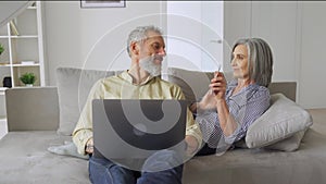 Happy mature older couple talking using laptop relaxing at home on couch.