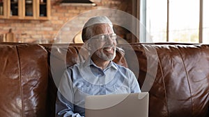 Happy mature senior 70s man using laptop, sitting on sofa