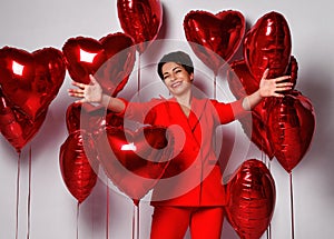 Happy mature short hair woman is posing with her arms spread surrounded by red heart air balloons. Holiday party