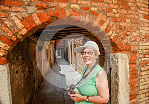 Happy mature tourist woman stays against narrow street in Venice