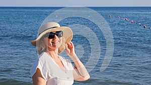 Happy mature woman 50 years old dressed in white dress, straw beach and sunglasses on the beach near the seashore