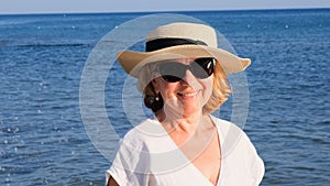 Happy mature woman 50 years old dressed in white dress, straw beach and sunglasses on the beach near the seashore