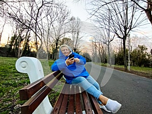 Happy mature woman in blue hoodie looking at mobile, sitting on bench in park at sunset.