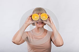 Happy mature woman covering her eyes with orange halves and smiling on light background. Vitamins for aging beauty