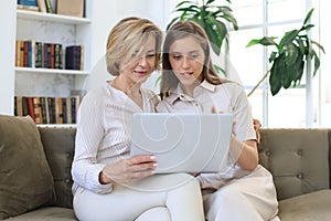 Happy mature woman with her adult daughter smiling while sitting on sofa and using laptop in living room