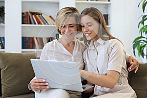 Happy mature woman with her adult daughter smiling while sitting on sofa and using laptop in living room