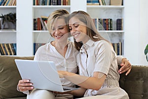 Happy mature woman with her adult daughter smiling while sitting on sofa and using laptop in living room