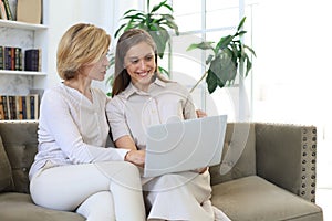 Happy mature woman with her adult daughter smiling while sitting on sofa and using laptop in living room