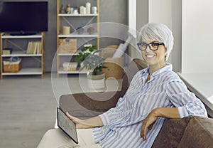 Happy mature woman sitting on sofa and planning her time using planner app on tablet