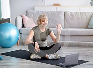 Happy mature woman in sportswear sitting on yoga mat in front of laptop, greeting her personal trainer before training