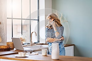 Happy mature woman standing in the kitchen at home and talking with somebody on her smartphone