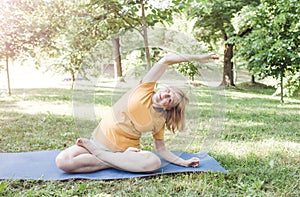 A happy mature woman in a yellow T-shirt is doing yoga sitting in the park in the lotus position doing sideways bends