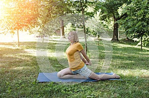 A happy mature woman in a yellow T-shirt is doing yoga sitting in the park in the lotus position doing sideways bends