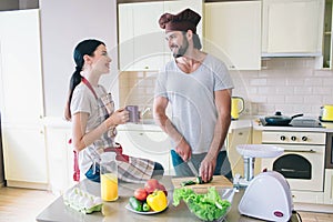 Happy man and woman stand in kitchen and look at each other. Guy cuts cucumber with knife. Young woman holds cup of tea