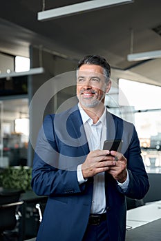 Happy mid aged ceo businessman wearing suit holding cell phone in office.