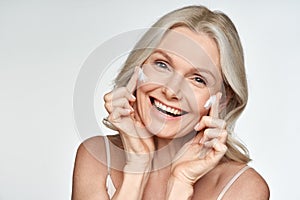 Happy mid aged woman applying cream on face skin isolated on white background.