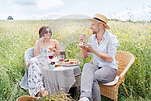 Happy middle age couple sitting in rattan chairs on the high grass meadow next the table served with fruit pie dish and vintage