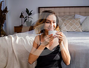 Happy middle-aged blond woman holding a cup of tea or coffee looking at the camera in cozy home
