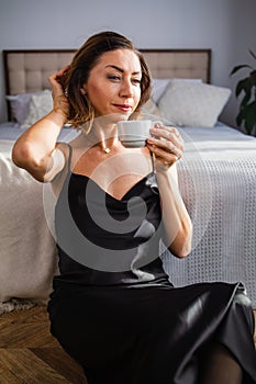 Happy middle-aged blond woman holding a cup of tea or coffee looking at the camera in cozy home