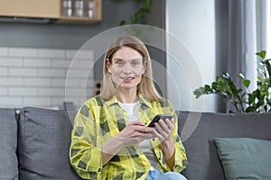 Happy middle-aged blonde woman sitting on sofa at home looking at camera and smiling holding phone in hands