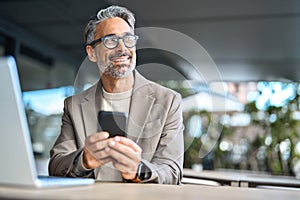 Happy middle aged business man using phone sitting outside office looking away.