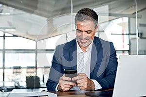 Happy middle aged business man wearing suit sitting in office using cell phone.