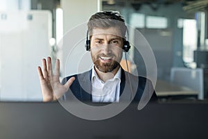 Happy middle aged businessman in headphones waving hand at computer webcamera, having meeting in office interior