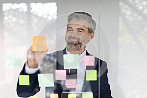 Happy middle aged businessman writing on sticky paper notes on glass wall