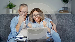 A happy middle-aged couple, looking at a laptop, feels like an online winner.