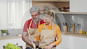 Happy middle aged couple in love cooking dinner together at kitchen, playful man embracing his beautiful wife