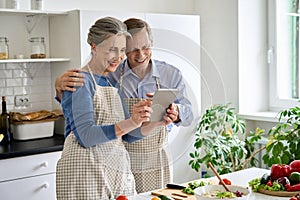 Happy middle aged couple preparing salad using tablet in kitchen.