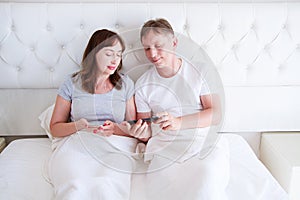 Happy middle aged couple smiling and chating on mobile phone in bedroom, smile people