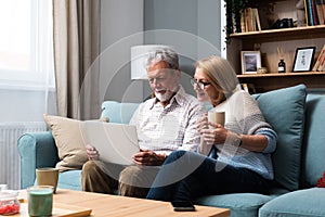 Happy middle aged family couple relaxing on sofa, using computer web surfing information shopping together at home. Elderly man