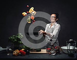 Happy middle-aged female chef tossing chopped vegetables in the air from a frying pan